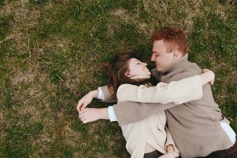 Cheerful couple outdoors, lying on grass, embracing warmly in cozy sweaters, relaxed pose, natural colors, soft breeze, candid romance, everyday happiness, intimate moment