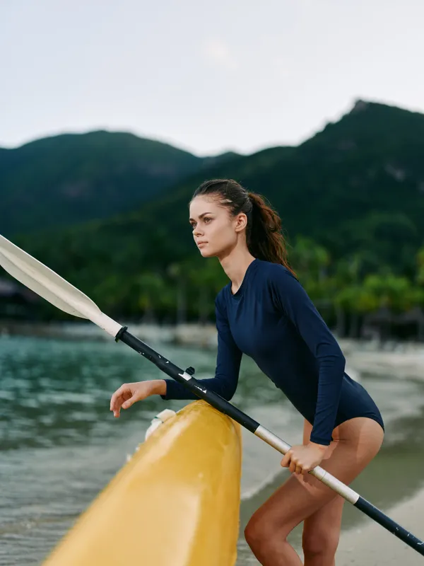 Female paddler in a navy wetsuit stands at the bow of a yellow paddleboard, gripping a paddle as calm water and distant mountains frame a tranquil coastal scene.