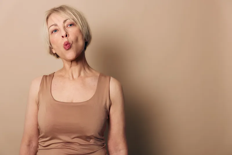 Mature woman with short blonde hair wearing beige tank top makes playful face on neutral background. Concept of aging confidence, individuality, and lighthearted expression.