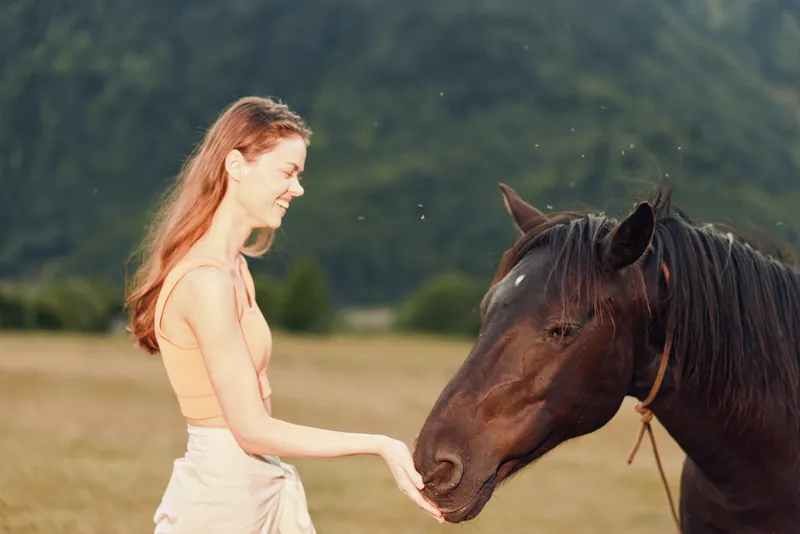 A smiling woman in a light peach dress gently interacts with a calm horse in an open meadow under soft sunlight, creating a tranquil rural moment filled with harmony.