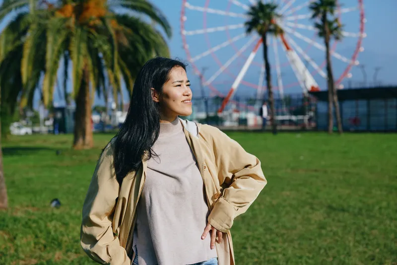 Woman in casual beige jacket and sweater stands in a sunny park with palm trees and a ferris wheel in the background, capturing carefree outdoor fashion and travel vibes