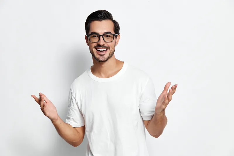 Happy cheerful young man wearing glasses and a white t-shirt smiling at the camera while gesturing with his hands, isolated on a plain white background.
