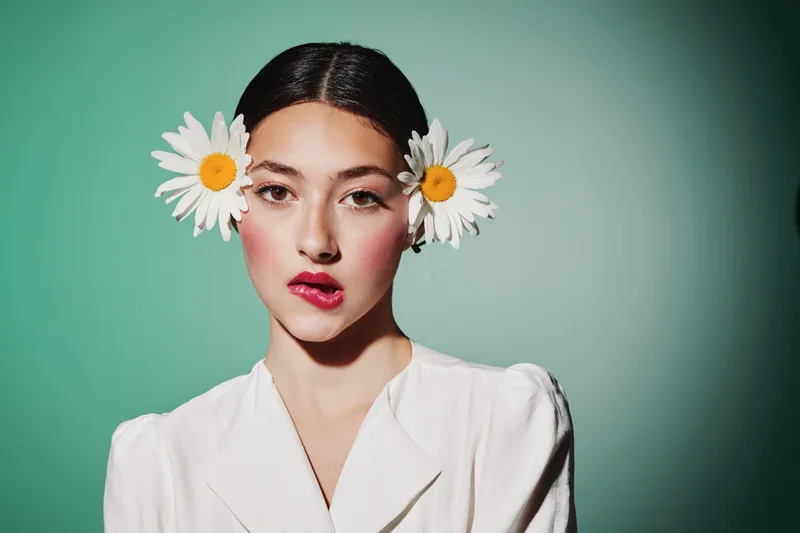 Young woman in white blouse with daisies in hair posing brightly on green background with soft makeup, lipstick, and creative beauty styling concept for fashion and expression.