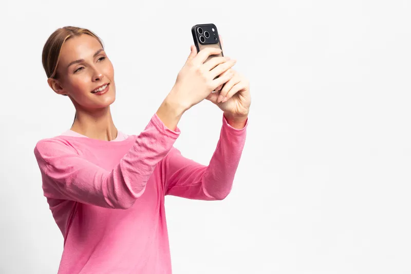 Young woman in pink sweater takes selfie with smartphone on white background. She smiles slightly with natural light illuminating her face, concept for social media, communication, and technology.