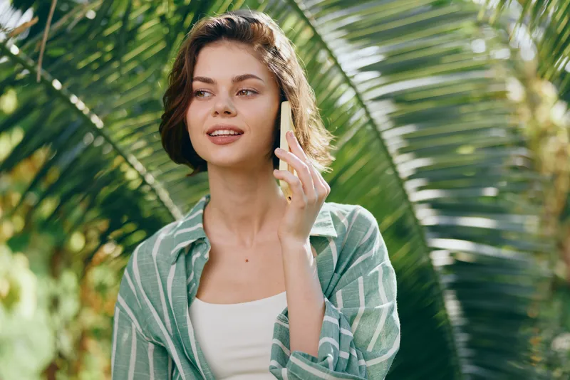 Portrait of a young woman speaking on a smartphone outdoors among lush tropical greenery, casual striped shirt, relaxed mood.