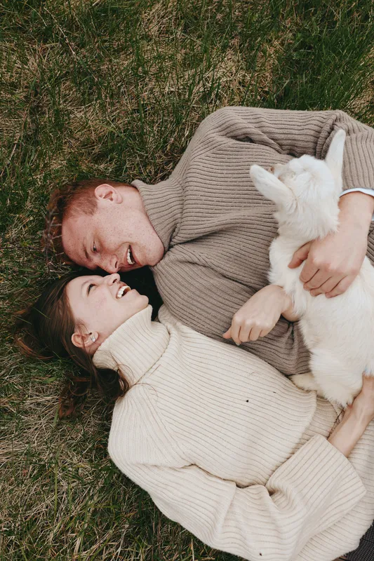 Goat, kid, couple, outdoor scene shows two people lying on grass, smiling, cuddle, affection, relaxation in a cozy rural lifestyle setting with a gentle farm vibe and playful animal presence