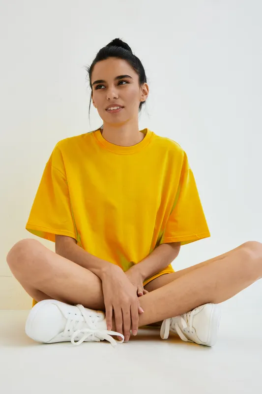 young woman sitting cross-legged on the floor in a bright yellow oversized t-shirt with white sneakers, casual pose against a plain white background, relaxed mood