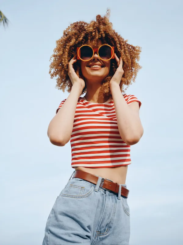 Portrait of a cheerful woman wearing a red striped crop top and round sunglasses, standing outdoors beneath a clear blue sky with warm sunlight and a carefree summer vibe.