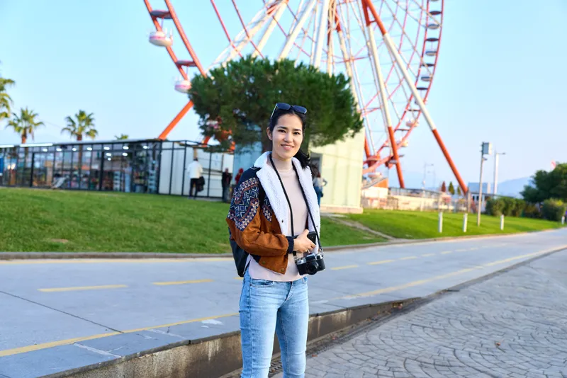 Camera in hand, traveler explores outdoor amusement park by a towering ferris wheel, casual fashion, confident stance, daytime scene, photography subject capturing joyful leisure and urban scenery