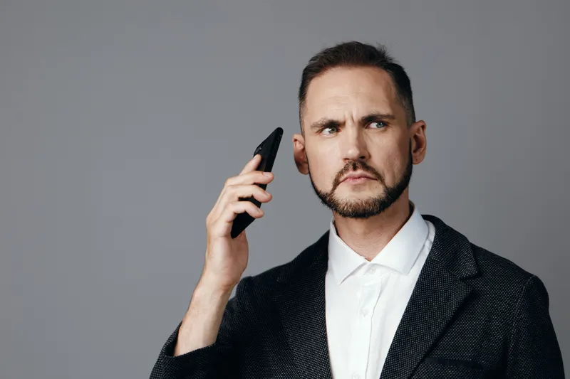 A focused business professional man stands against a solid color background, exuding confidence and intent while he speaks on a mobile device.