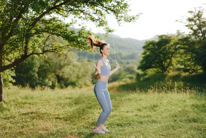 Young athletic woman jumping rope outdoors in a vibrant green field, embracing fitness with joy and energy against a scenic nature backdrop