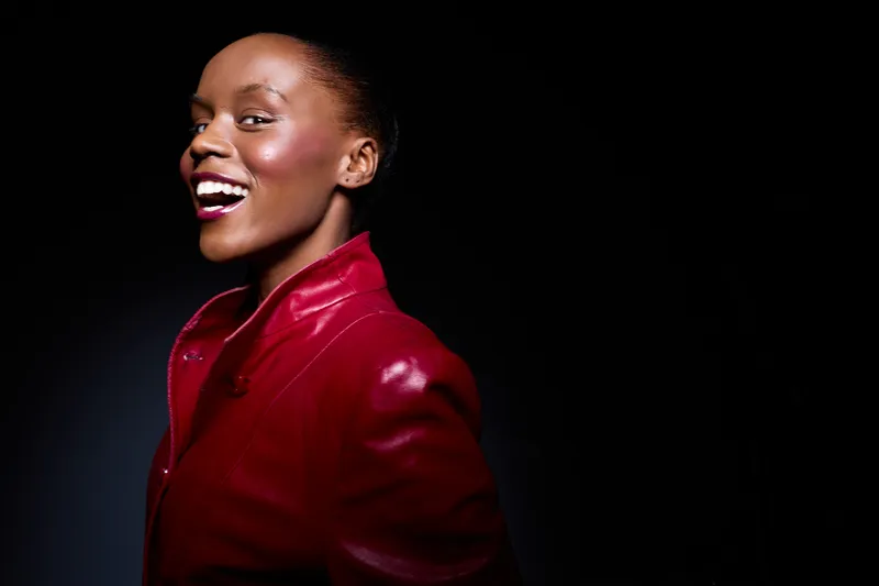 Smiling African woman with dark skin wearing a red leather jacket poses against black background. Studio portrait with joyful expression and stylish fashion concept for beauty and confidence.