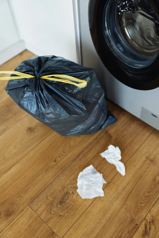 Washing machine, trash bag, laundry and floor in a domestic utility room with tied black garbage sack and crumpled tissues on wooden floor beside appliance for household cleaning.