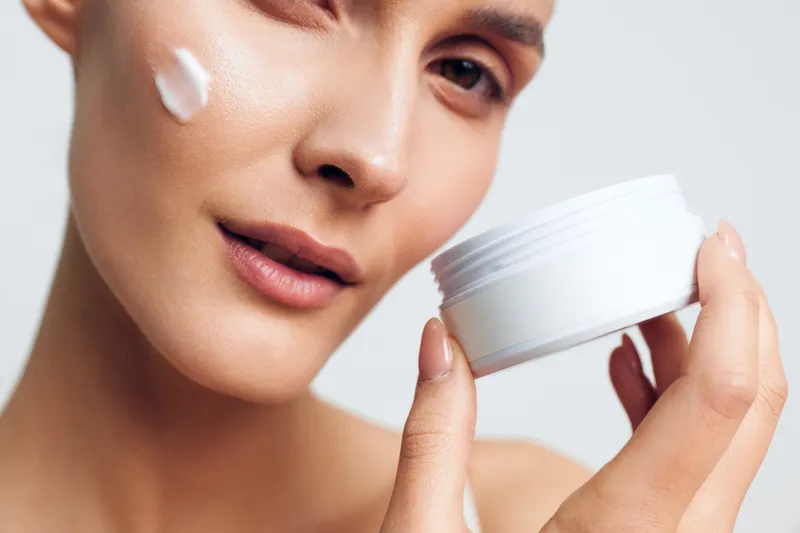 A close up of a woman applying white cream to her cheek while holding a white jar. The image represents skincare, beauty routine, and healthy skin care concepts.