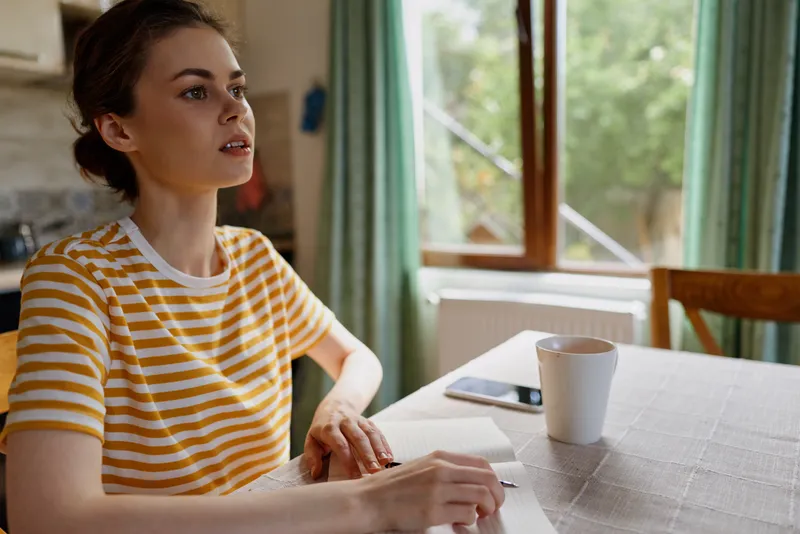 Woman with short hair wearing a striped shirt sits at a table near a window, writing in a notebook beside a mug. Daylight fills a calm, cozy home scene with relaxed focus.