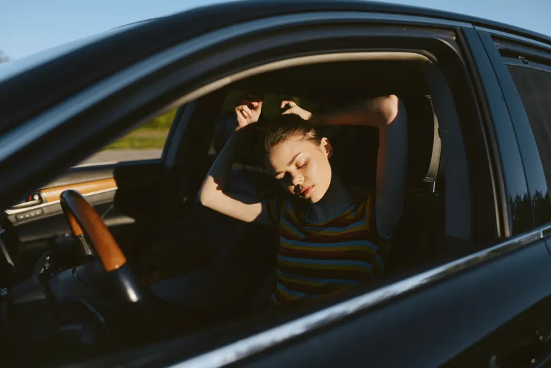 Candid woman inside a car adjusting hair, sunlit moment, casual lifestyle, retro film color tones, warm shadows, relaxed mood, everyday travel scene