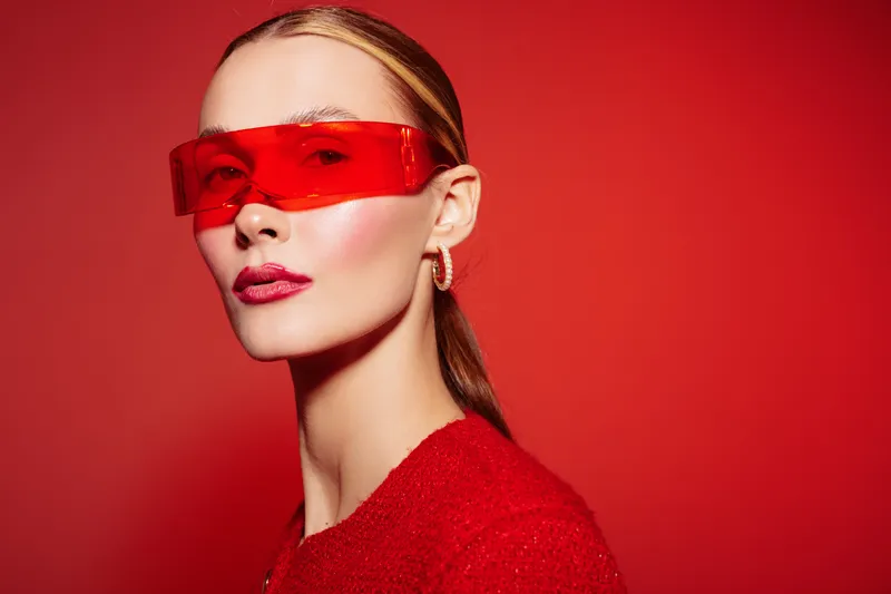 Stylish woman with slicked hair wearing red transparent glasses and red outfit poses confidently on red background. Concept of fashion, modern style, and bold accessories.