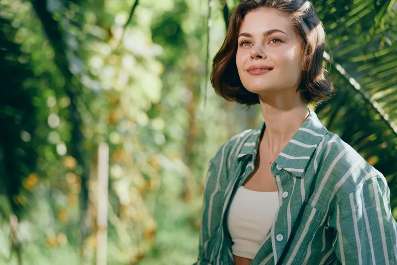 Portrait of a young woman outdoors, smiling softly in a striped green shirt, sunlit garden setting and natural breeze.