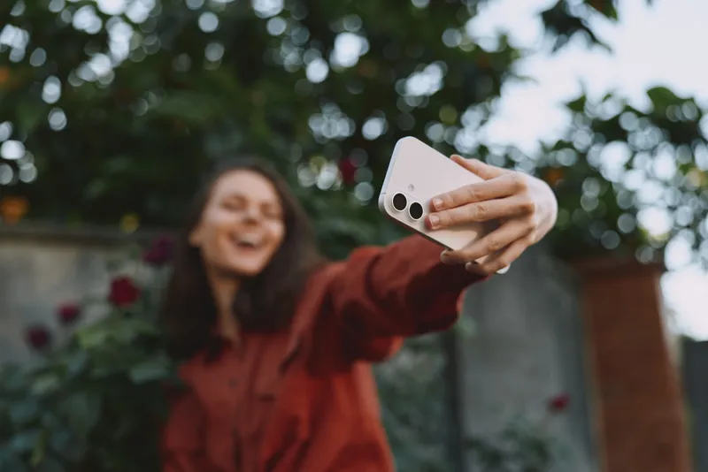 Smiling woman taking a selfie outdoors with a smartphone, dressed in a red jacket. Casual lifestyle shot highlighting connection, fun, and self expression in daylight