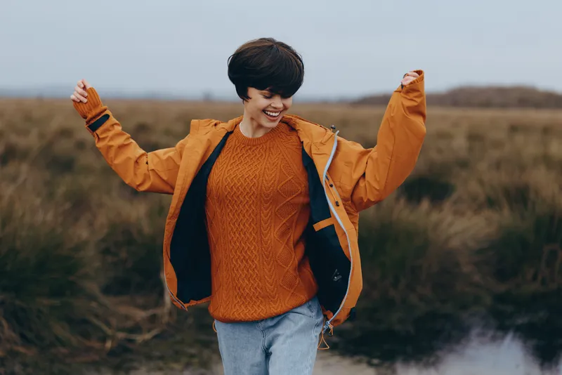 Woman lifestyle outdoors field in autumn, orange jacket and sweater, smiling with arms open, casual travel mood, wind in hair, soft countryside background golden hour and distant clouds