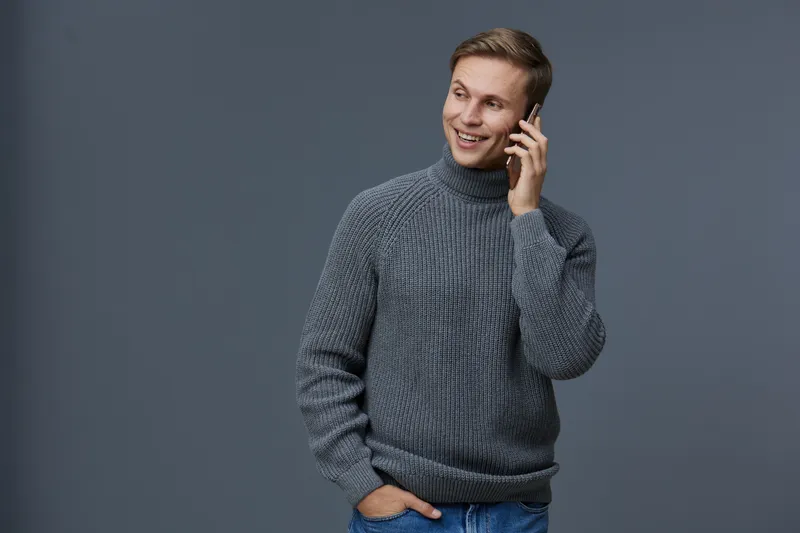 Male smiling man talking on mobile phone, casual wear gray knit sweater, modern studio setting, man using smartphone, cheerful expression, technology, lifestyle, communication, positive mood, indoor s