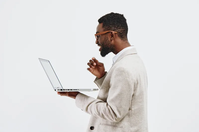 Professional young African-American man in smart casual attire holding a laptop, looking at the screen with focus isolated on plain white background. Business, technology, work, education, modern life
