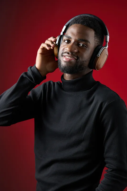 Young Black man enjoying music with headphones, smiling against a red background, stylish black turtleneck, showcasing a joyful and relaxed atmosphere