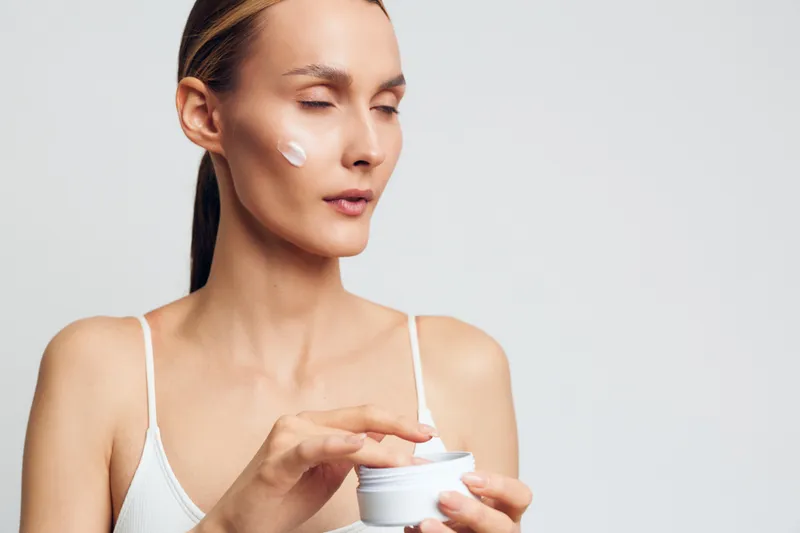 Woman skincare close up applying face cream from jar for self care and wellness. Soft studio lighting on clean background supports calm skin hydration and beauty routine.