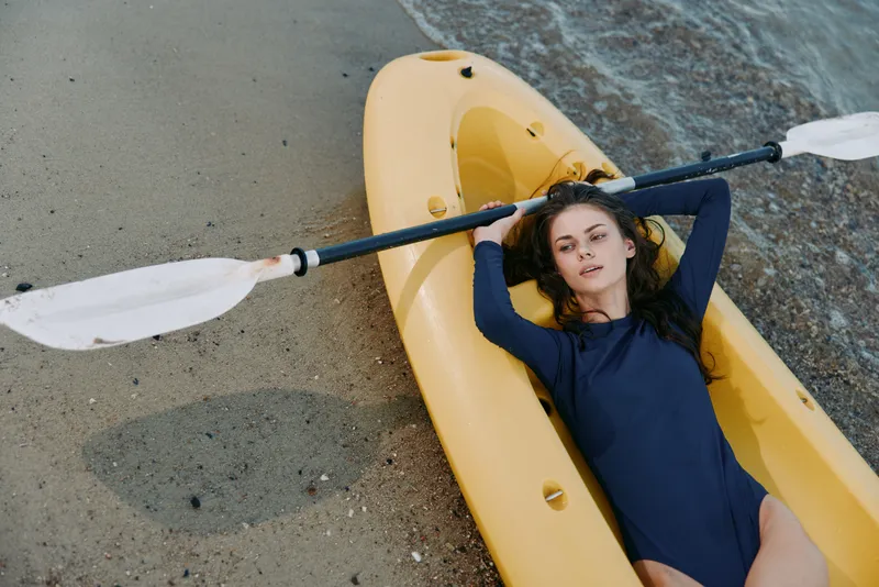 A young woman lies in a bright yellow kayak on a sandy shoreline, gripping a double blades paddle across her chest, resting after a calm beach paddle session.