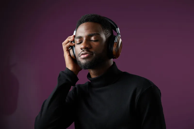 A young Black man enjoying music with headphones in a stylish black turtleneck against a purple background, showcasing emotions of relaxation and contentment