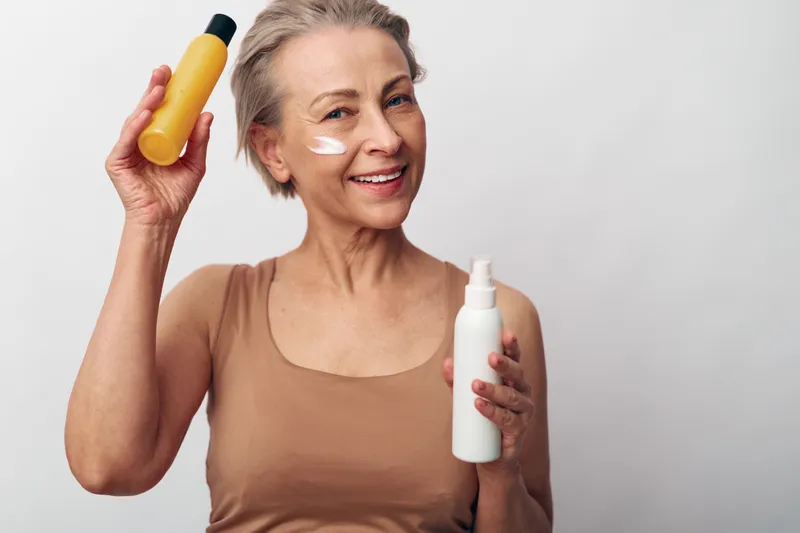 Smiling mature woman with short hair holds yellow bottle and white spray container, applying cream on cheek. Concept of skincare, beauty routine, anti aging and self care at home.