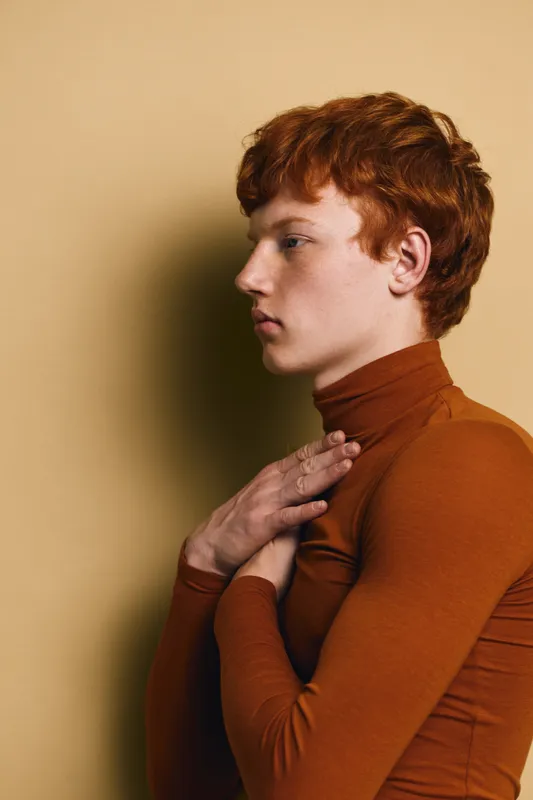 Young redhead person in brown turtleneck with clasped hands near chest, thoughtful profile portrait with calm expression on beige background. Side view, studio shot of pensive youth.