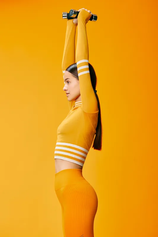 Sporty woman fitness portrait: fit female in bright yellow activewear lifting small dumbbells overhead, side profile with sleek ponytail, focused determined expression, minimalist monochrome orange