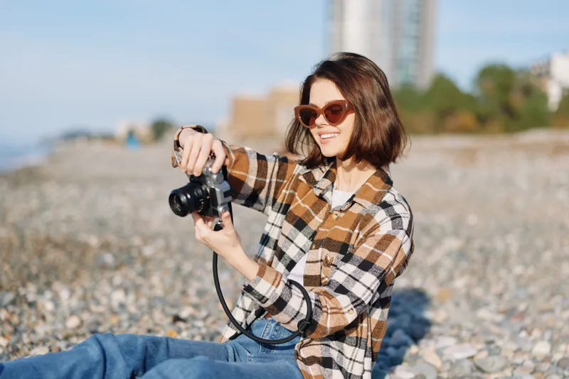 Photographer, woman, camera, outdoor, beach, casual, sunglasses, plaid shirt, shooting, lifestyle in sunny scene capturing candid coastal moments with relaxed pose and blue jeans