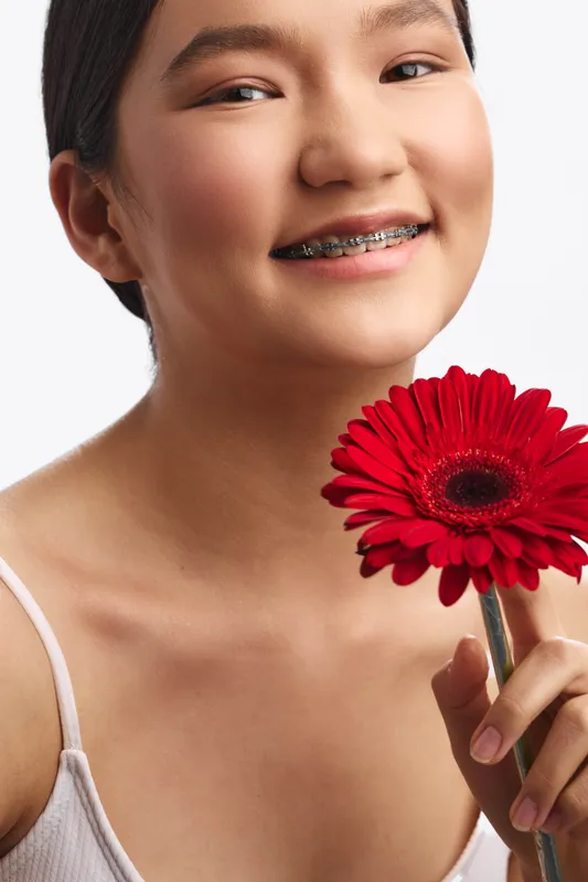 Asian woman smiles softly, holding a bright red flower near her face in a studio portrait. Warm light highlights calm confidence and gentle charm.