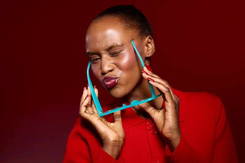 Portrait of a smiling African woman with dark skin holding turquoise sunglasses near her face against a red background. Concept of fashion, beauty, and joyful expression.