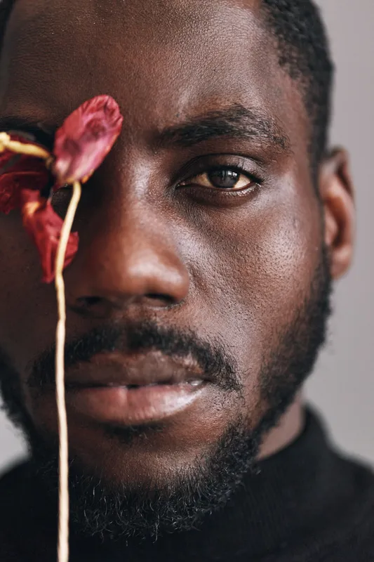 African man with dark skin and beard in creative conceptual style closeup portrait holding dried flower near face with serious expression and natural lighting on neutral background.