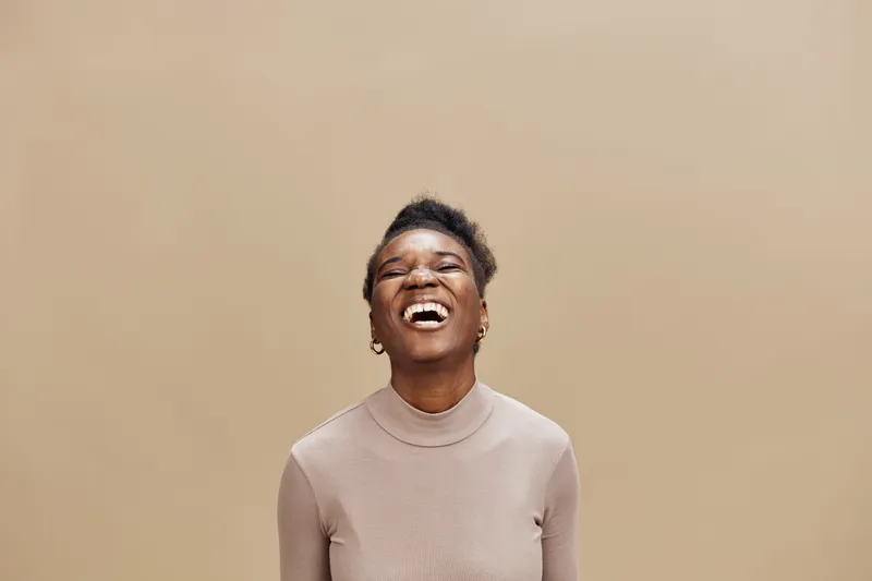 Joyful black woman laughing against a beige background wearing a light sweater, conveying happiness and positivity through her vibrant expression and relaxed style
