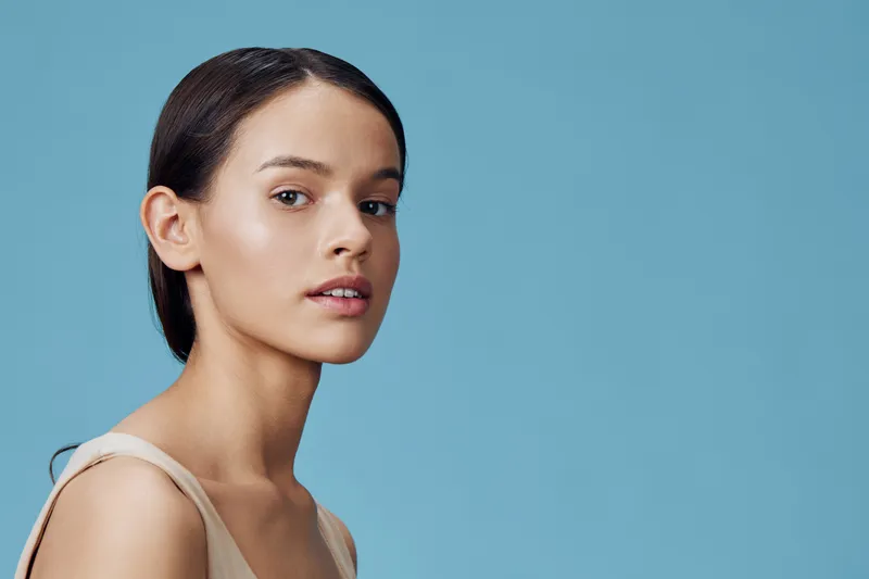 Beautiful young model with smooth skin and minimal makeup against a soft blue background, showcasing natural beauty and elegance in a serene expression
