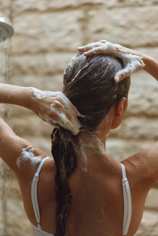 Woman washing hair under shower with soap foam on textured wall background, showing wet dark hair and skin care in natural light and calm mood.