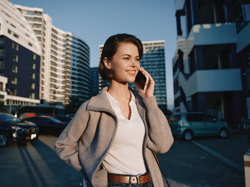 A cheerful professional woman talks on a mobile phone in a modern urban setting, smiling and confident as she walks through a sunny city street.