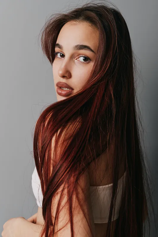 Woman with long brown hair and natural makeup posing in white top against gray background for beauty, fashion, and portrait photography concept with soft lighting and casual style.
