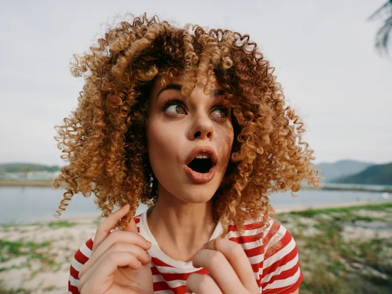 A surprised woman with voluminous curly hair stands at a coastal shoreline, eyes wide, mouth open in astonishment, striped shirt under natural daylight.