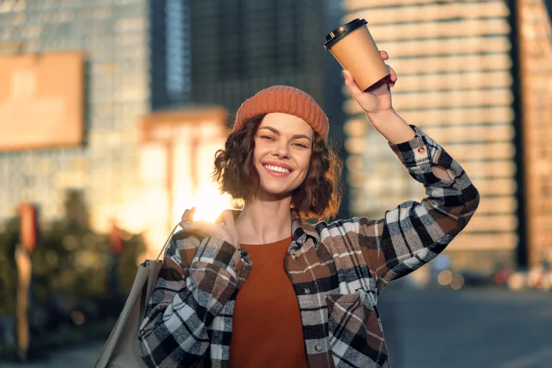 Woman coffee city smile beanie tote sunlight urban: young woman raises a takeaway cup on a sunlit street, candid lifestyle portrait with golden hour glow, authenticity and mindful living.