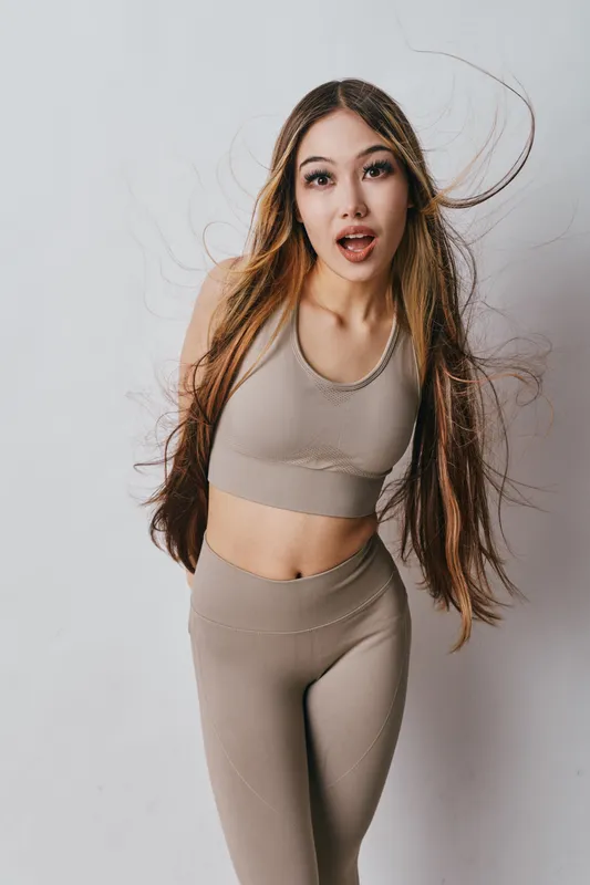 Young woman with long hair wearing beige activewear posing against white background. Fitness girl with open mouth expression in studio for health and lifestyle.