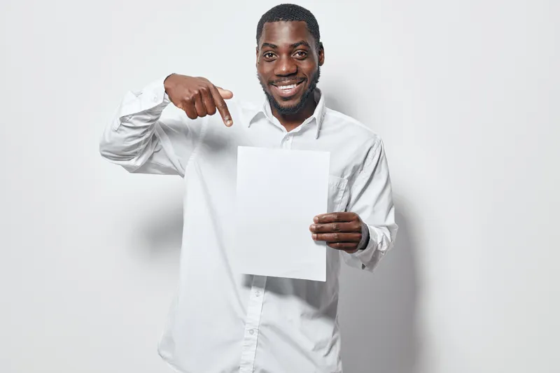 African American man with dark skin wearing white shirt smiling and pointing at blank paper on white background for presentation or advertisement concept.