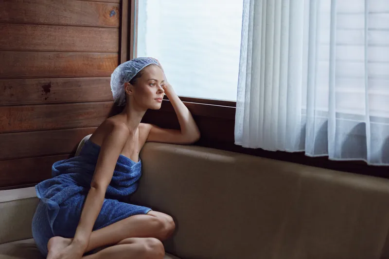 Woman wrapped in blue towel sitting indoors by the window, looking thoughtful with soft natural light and wooden wall background in cozy atmosphere.