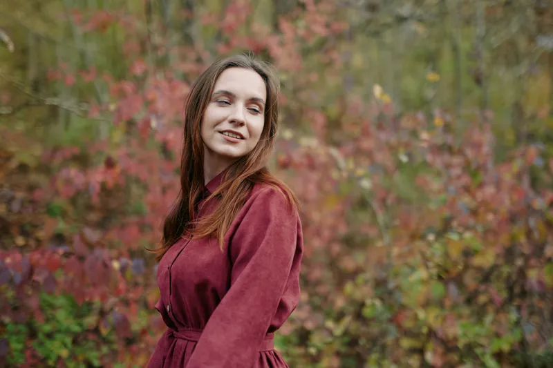 A candid autumn portrait of a young woman in a burgundy coat amid colorful foliage, conveying authentic emotion and realistic presence with natural expressions and gentle eye contact.