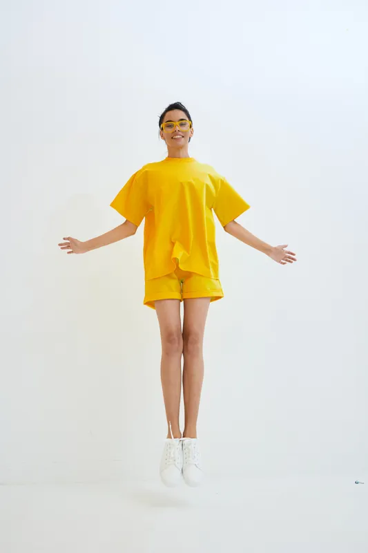 Young woman jumping in bright yellow casual outfit and white sneakers, smiling with arms outstretched against a clean white background, expressing joy and energy.