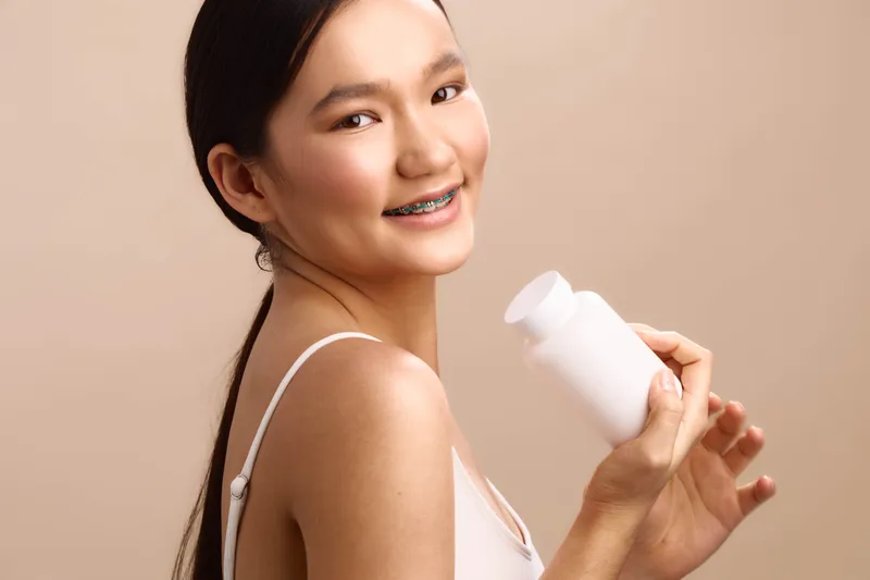 Asian woman smiles while turning toward the camera, holding a white bottle in a clean studio setting, conveying health, wellness, and a confident, fresh lifestyle.